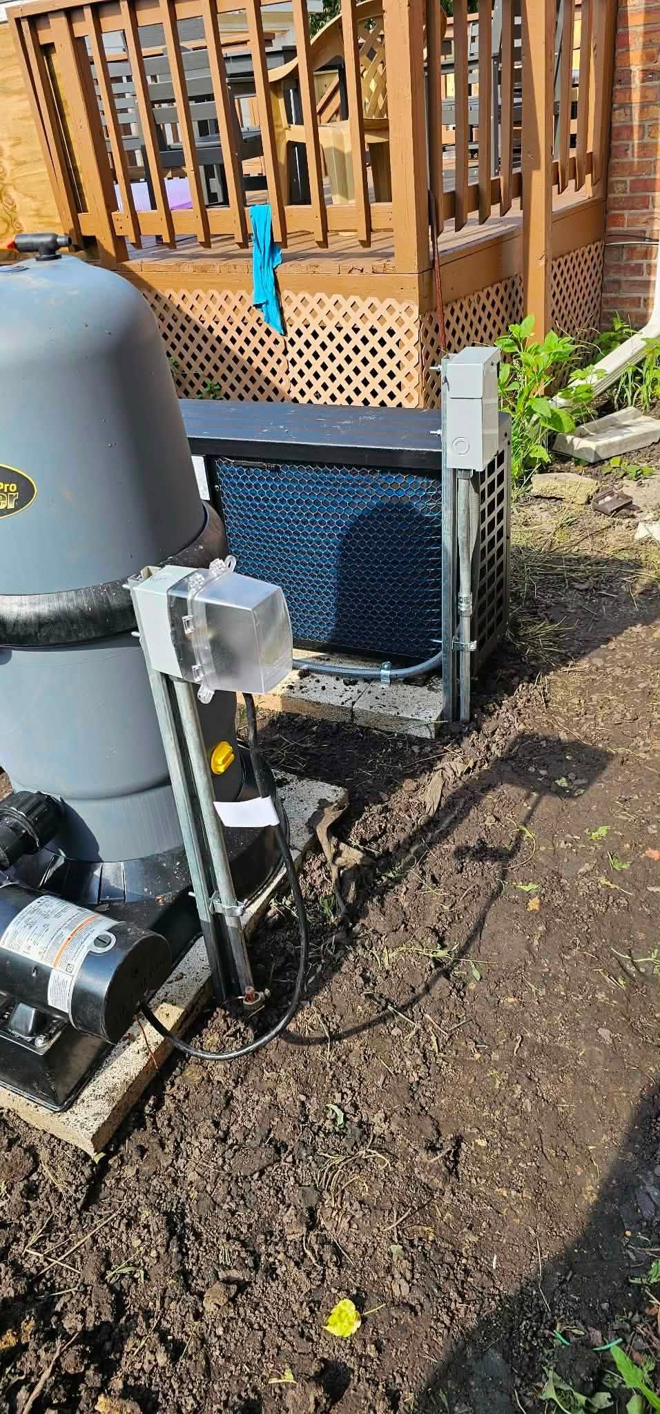 Outdoor utility equipment beside a wooden deck, with a gray tank, pipes, and a black grid cover