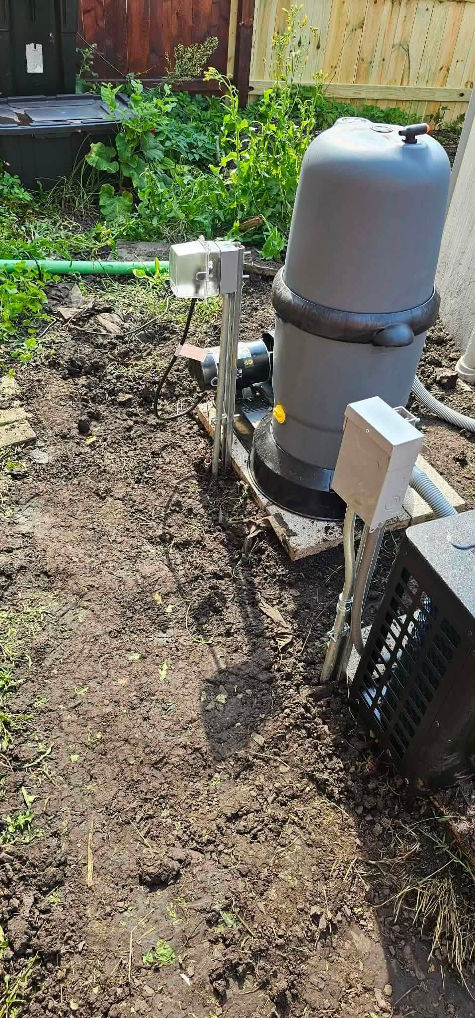 Backyard utility equipment with a gray tank, pipes, and a black vent on dirt and mulch.