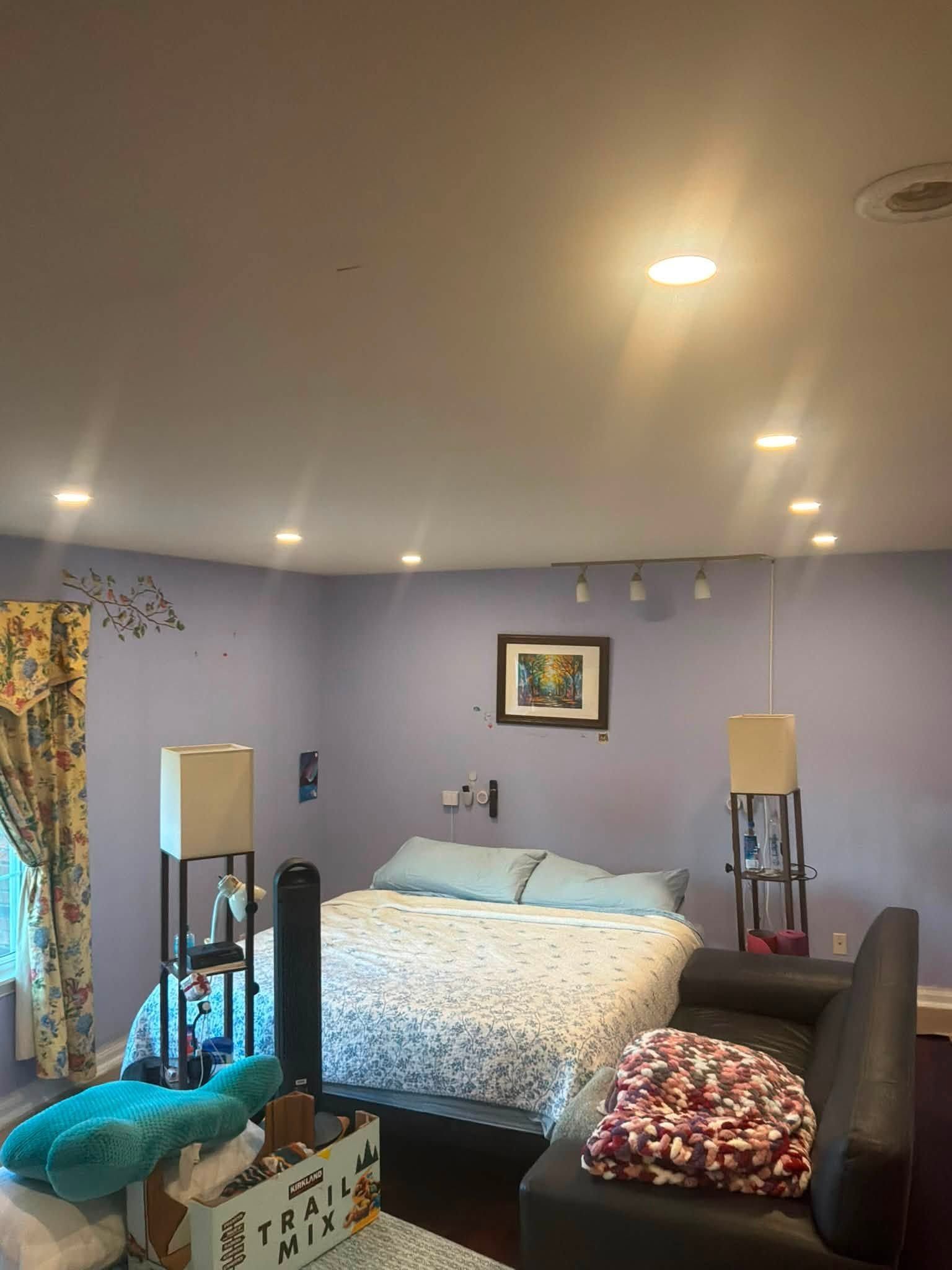 Bedroom with a bed, dark chairs, lamps, and lavender walls under recessed ceiling lights.