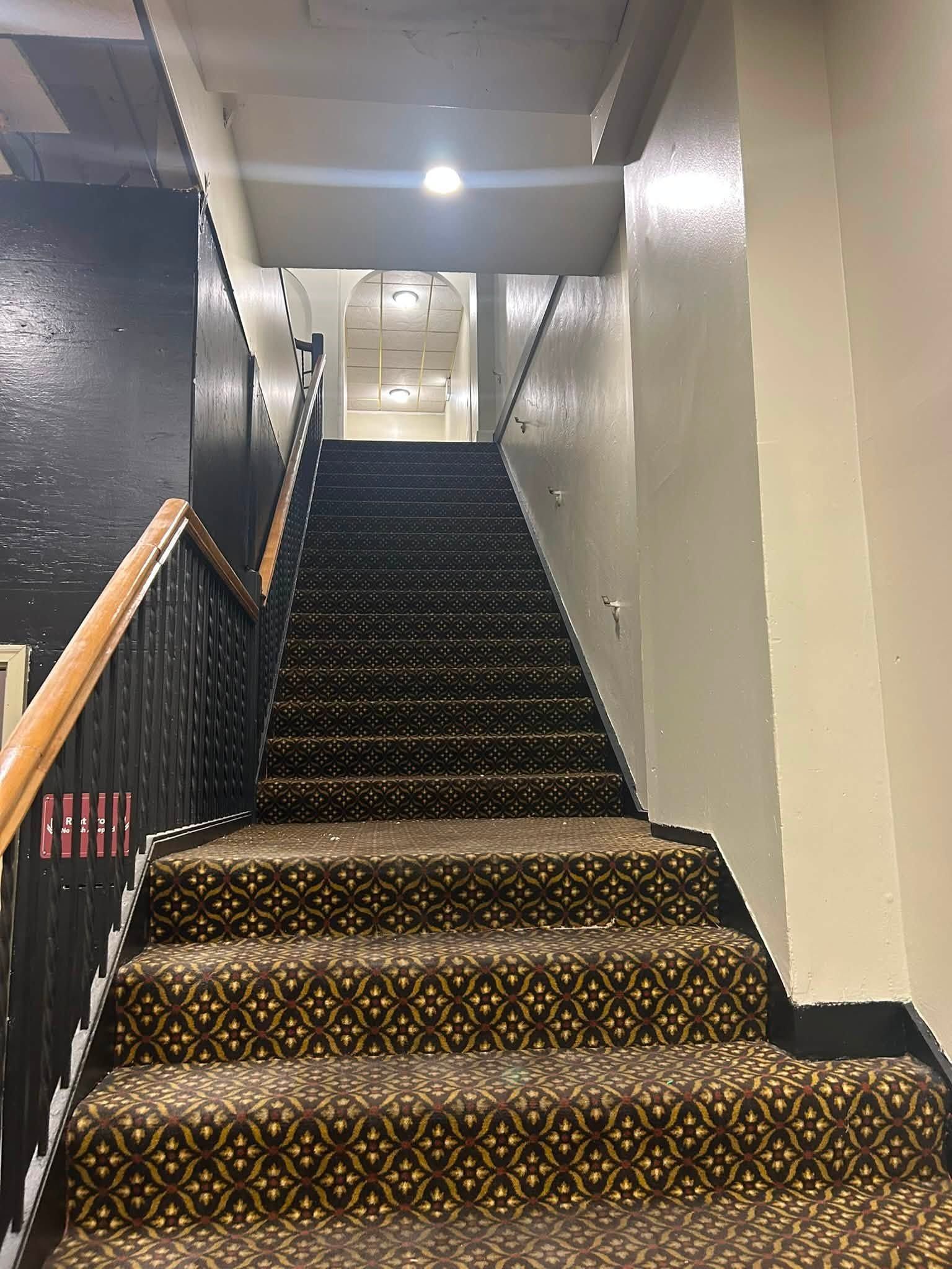 Staircase in an indoor hallway with patterned carpet and white walls, lit by ceiling lights.