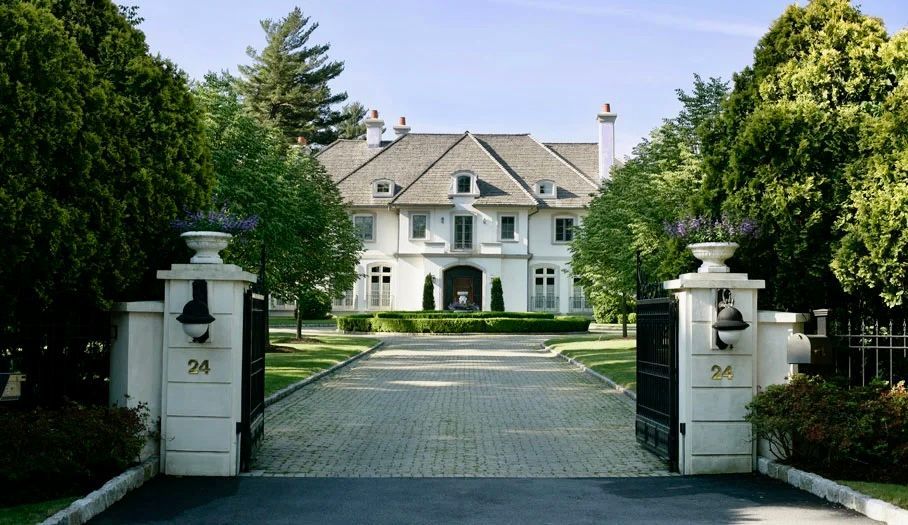 Large white mansion with a long driveway and black gate. Trees and manicured lawn.