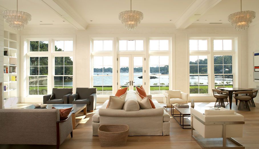 Bright living room with large windows overlooking a body of water. Light wood floors, white walls, and multiple seating areas.