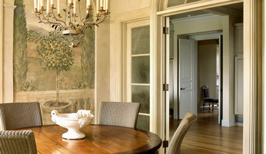 Dining room with round wooden table, chairs, mural, chandelier, and open doorway.