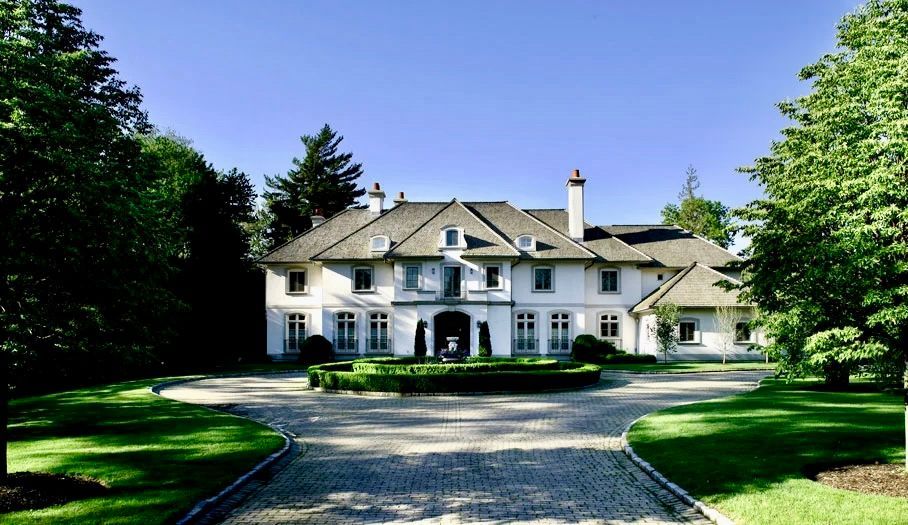White mansion with gray roof and cobblestone driveway, surrounded by greenery under a blue sky.