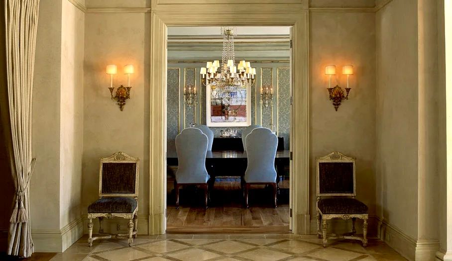 Elegant foyer with two ornate chairs, sconces, and view into a dining room with a chandelier.