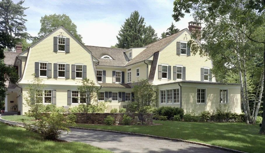 Yellow house with multiple dormers, green lawn, trees, and a long driveway.