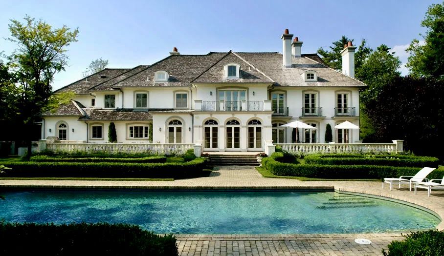 White mansion with a pool, brick patio, and manicured hedges, under a sunny sky.