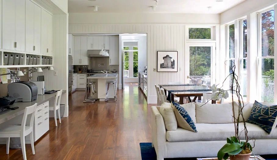 Spacious, bright living area with kitchen and home office visible. Hardwood floors, white walls, and large windows.