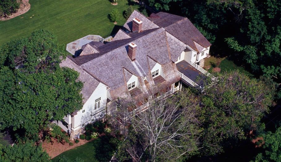 Aerial view of a large, two-story house with a brown shingle roof, surrounded by trees.