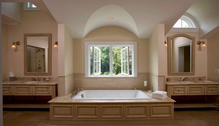 Spacious beige bathroom with a built-in tub, two vanities, and a large window overlooking trees.