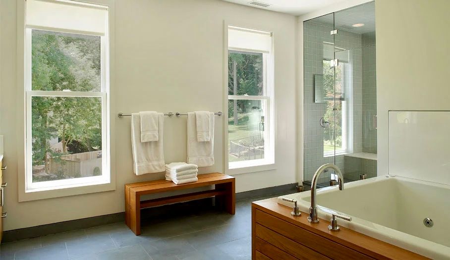 Bathroom with two windows, wood bench with towels, and a bathtub with a wood panel.