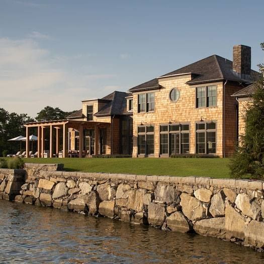 Large waterfront house with wood siding, a rock wall, and a green lawn.