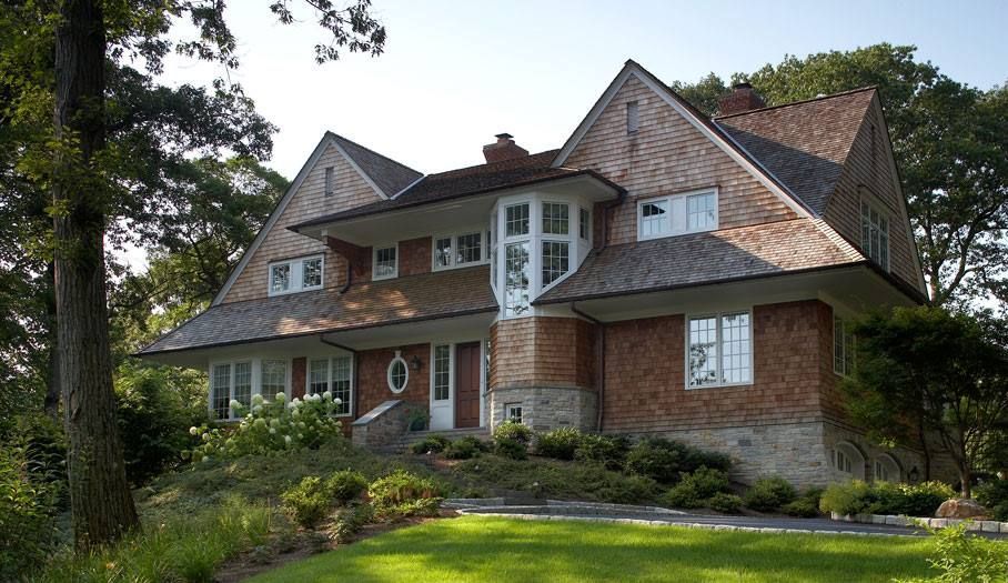 Brown brick and wood shingle house with gabled roof, set on a green lawn, surrounded by trees.