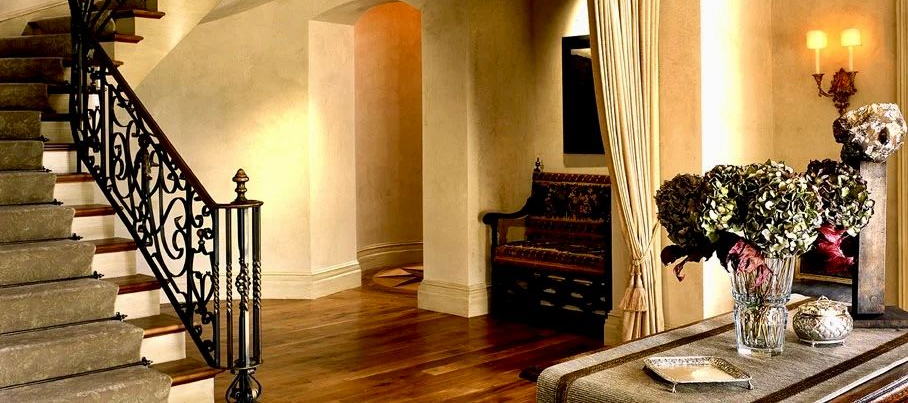Elegant foyer with a curving staircase, ornate iron railing, wooden bench, and floral arrangement on a side table.