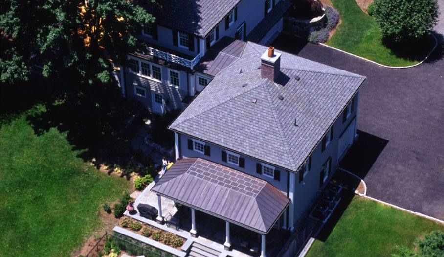 Aerial view of a gray house with a brown porch and dark roof, set on a green lawn with a black driveway.