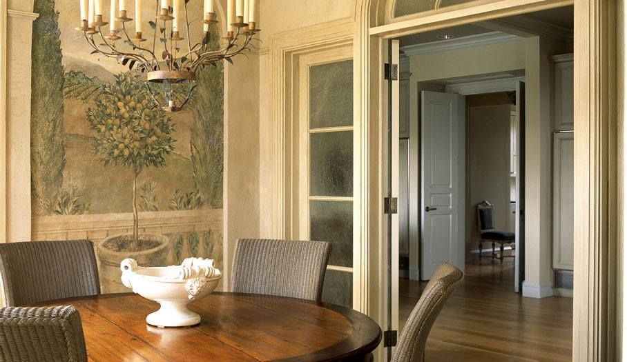 Dining room with mural, round table, chairs, and chandelier; open doorway to another room.