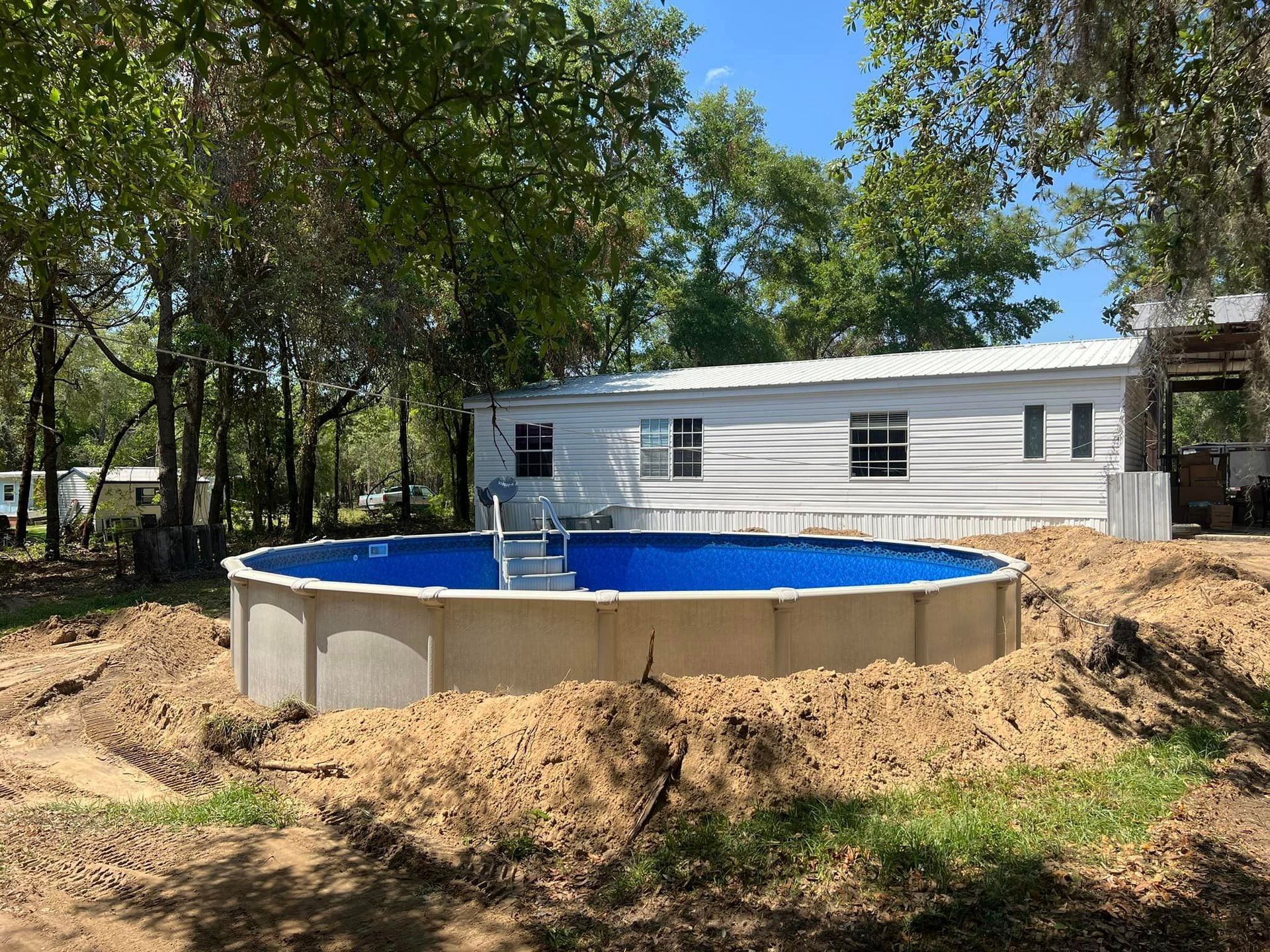 A large swimming pool is sitting in the dirt in front of a mobile home.