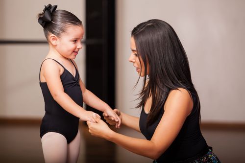 Coach helping a young gymnast stand in a studio, both smiling and holding hands.