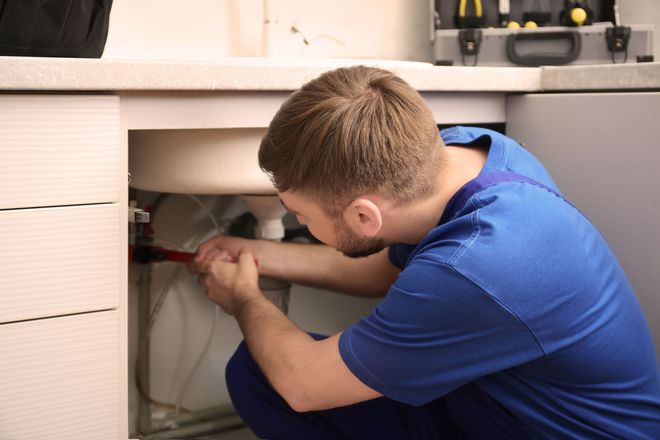 Technician in blue shirt kneels under a cabinet, tightening plumbing or wiring with a tool