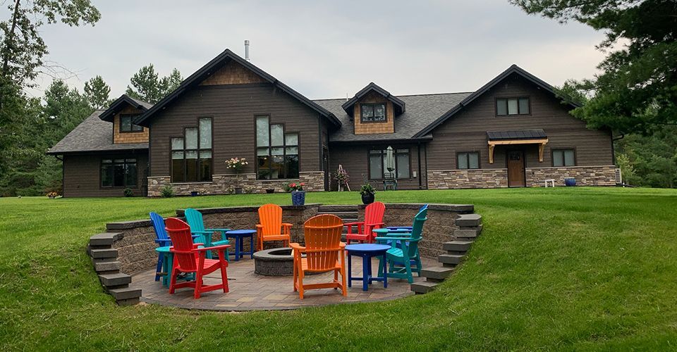 A group of colorful chairs are sitting around a fire pit in front of a large house.