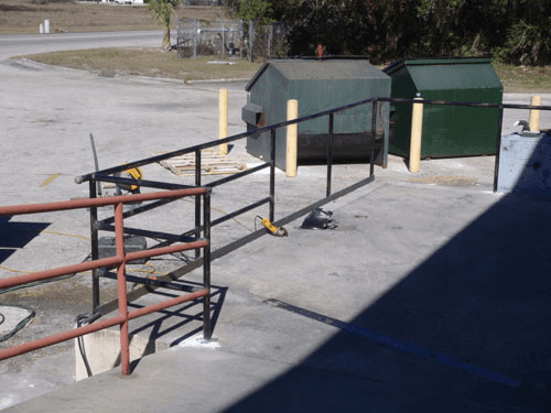 A metal railing in a parking lot with a green dumpster in the background