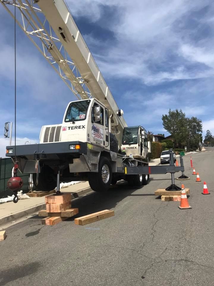 Crane truck on a sloped street, supported by wood blocks. Orange traffic cones mark the perimeter.