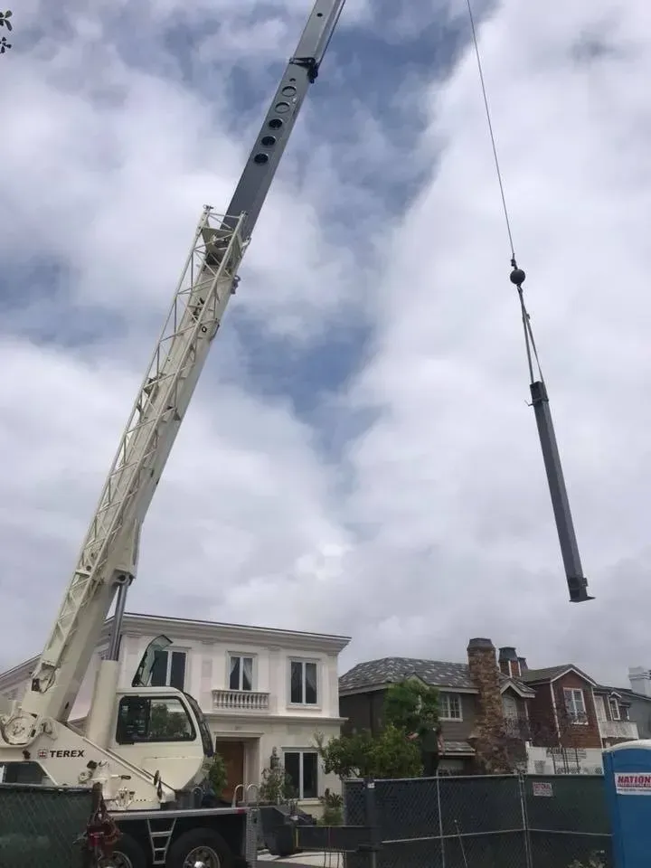 A large crane lifting a long, cylindrical object near houses under a cloudy sky.