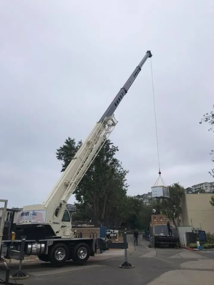 Crane lifting a rectangular object in a paved area, trees and building in the background under a cloudy sky.