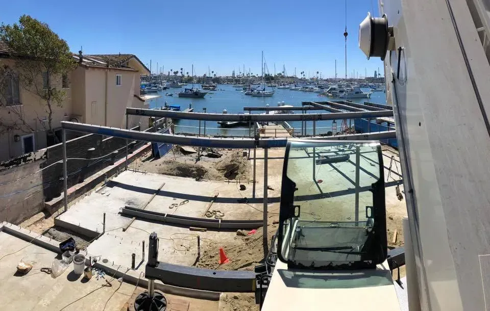 Construction site with harbor view: a crane, exposed concrete, and boats in the background.