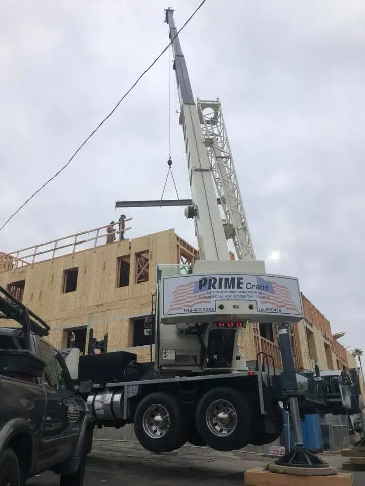 Crane lifting a steel beam onto a wood-framed building under construction on a cloudy day.