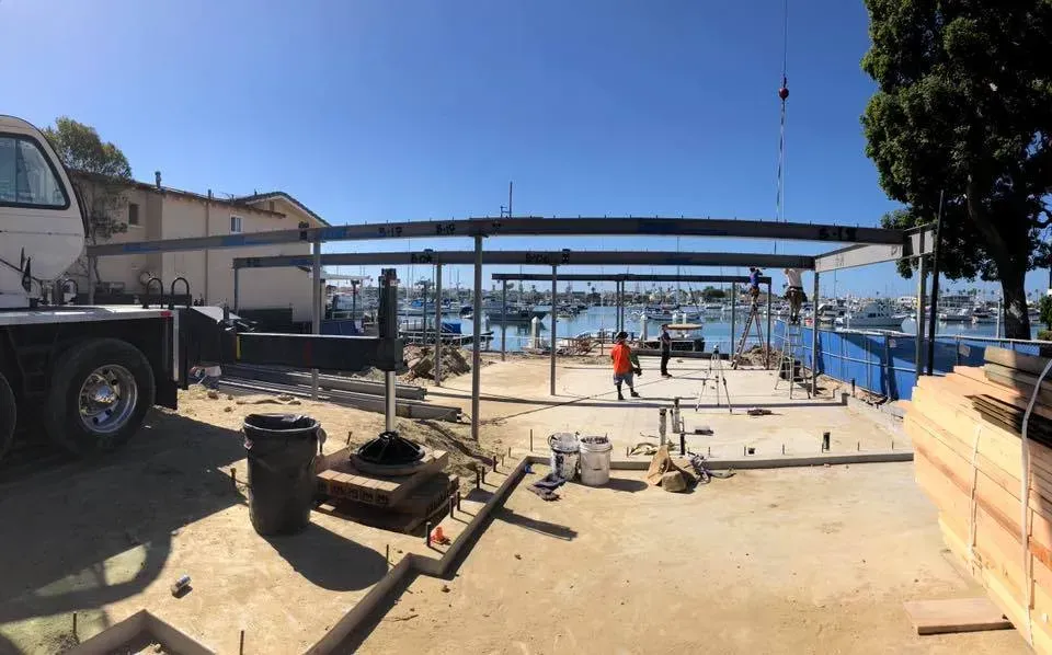 Construction site with steel beams, concrete, and workers, overlooking a harbor under a clear blue sky.