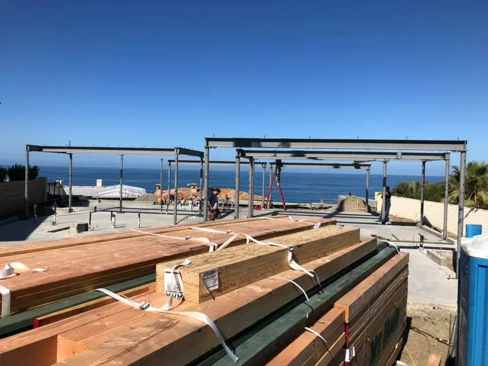 Construction site with steel framing overlooking ocean; lumber stacked in foreground.