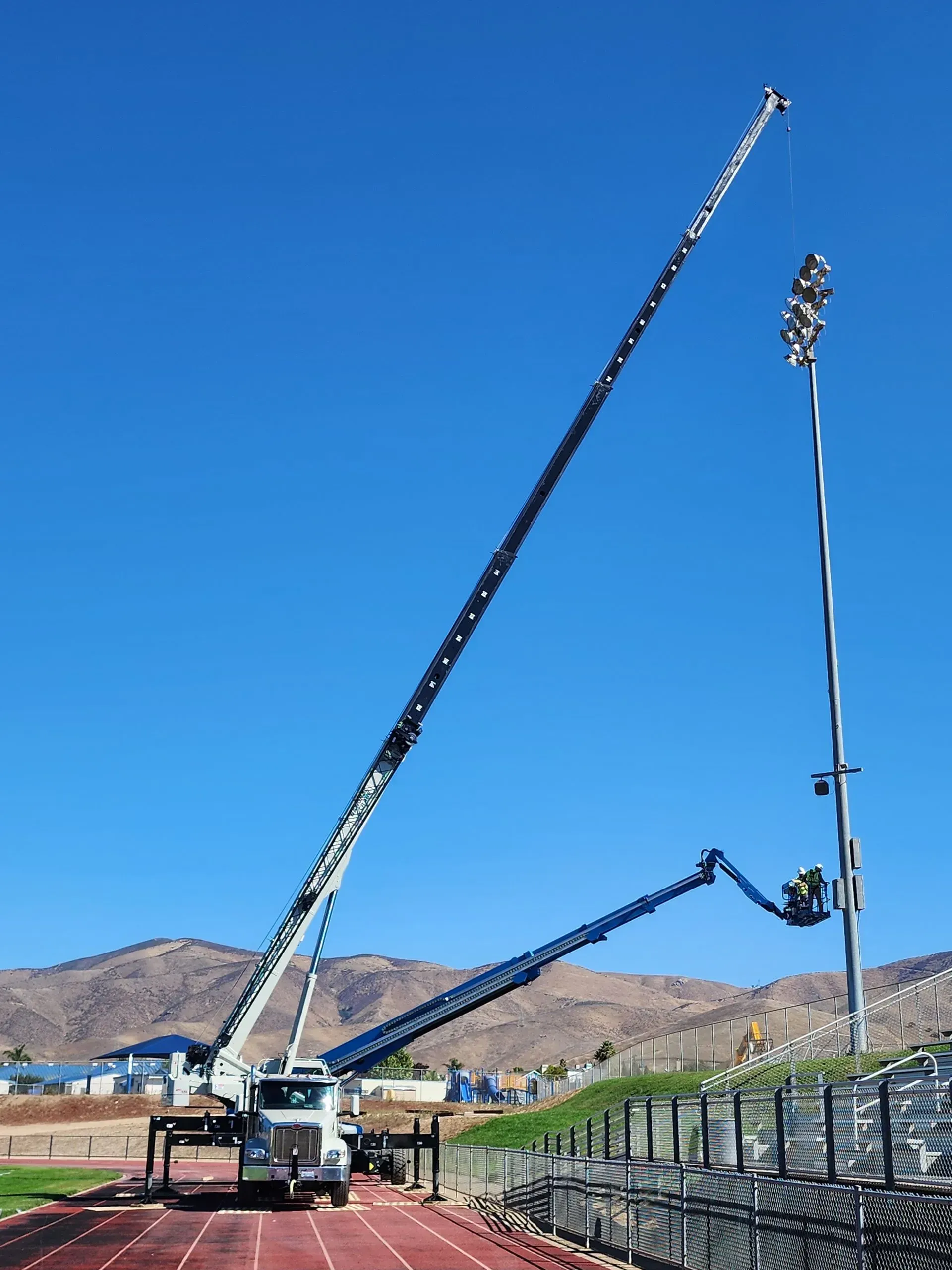 Truck-mounted aerial lift repairing stadium lights with a worker, track, blue sky.