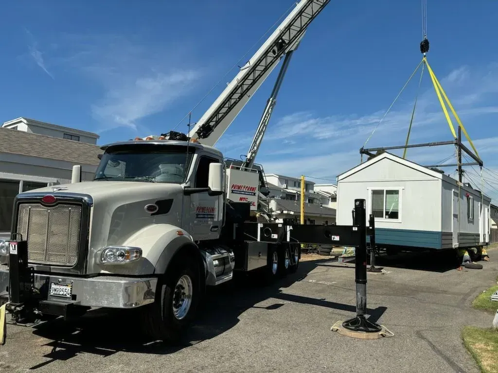 Crane lifting a small, white building onto a lot, bright sky overhead.