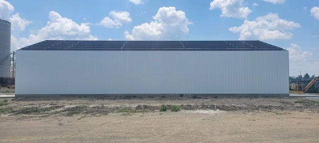 A large white building with a black roof is sitting in the middle of a dirt field.