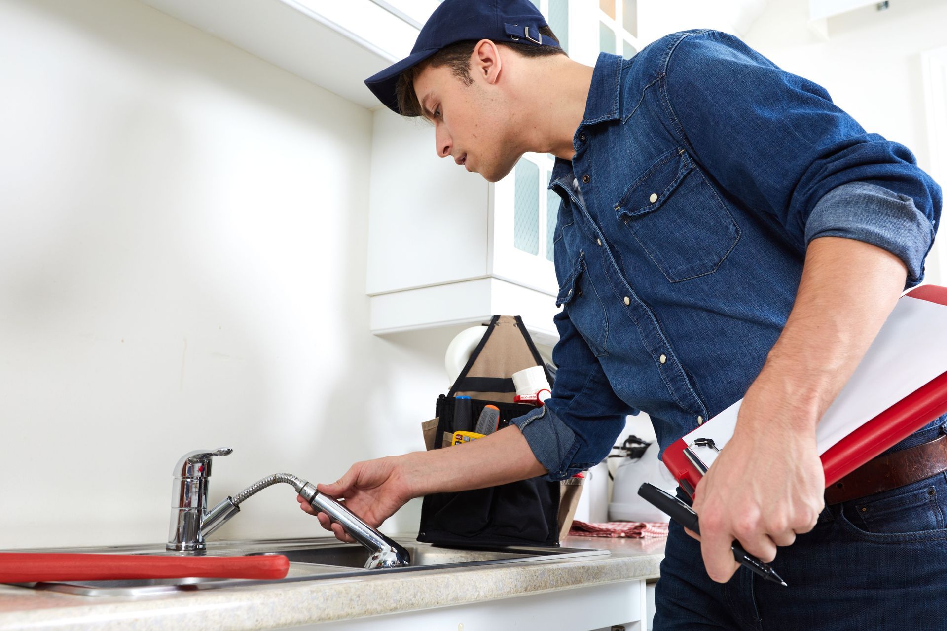 Plumber inspecting kitchen sink, wearing blue denim shirt and cap, holding clipboard and tools.