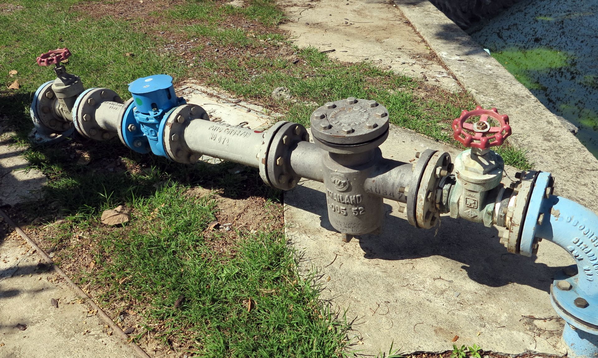 Industrial water pipe with blue and red valves running through a grassy concrete area