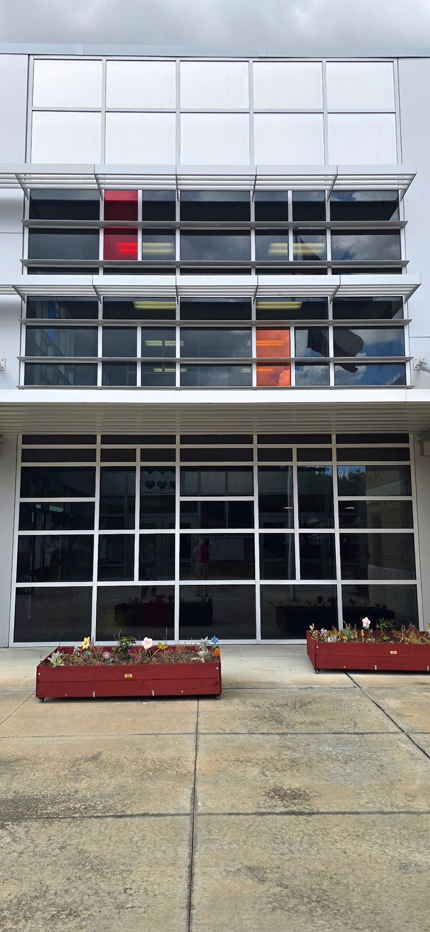 A modern two-story building facade featuring grid-pattern windows, red accents, and two rectangular planters in front.