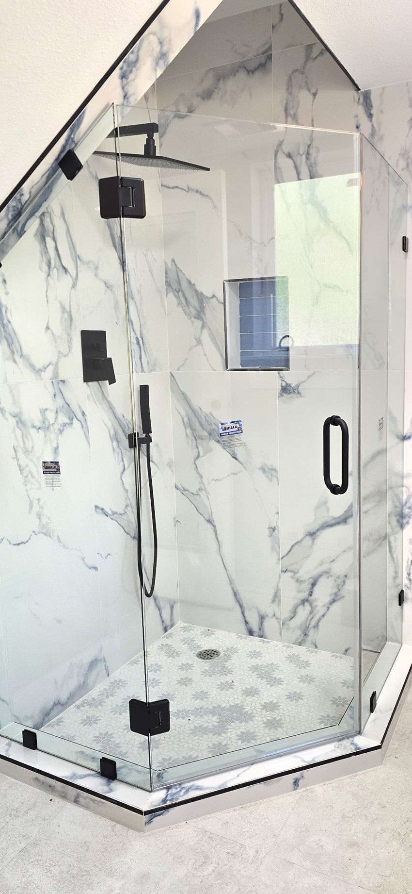 A modern walk-in shower with white and grey marble tile walls, a glass enclosure, black hardware, and a pebble floor.