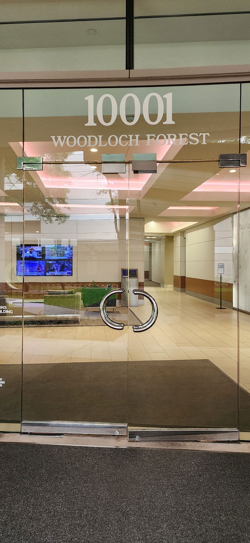 Glass entrance doors to the building at 10001 Woodloch Forest, featuring a circular logo and a view into the lobby.