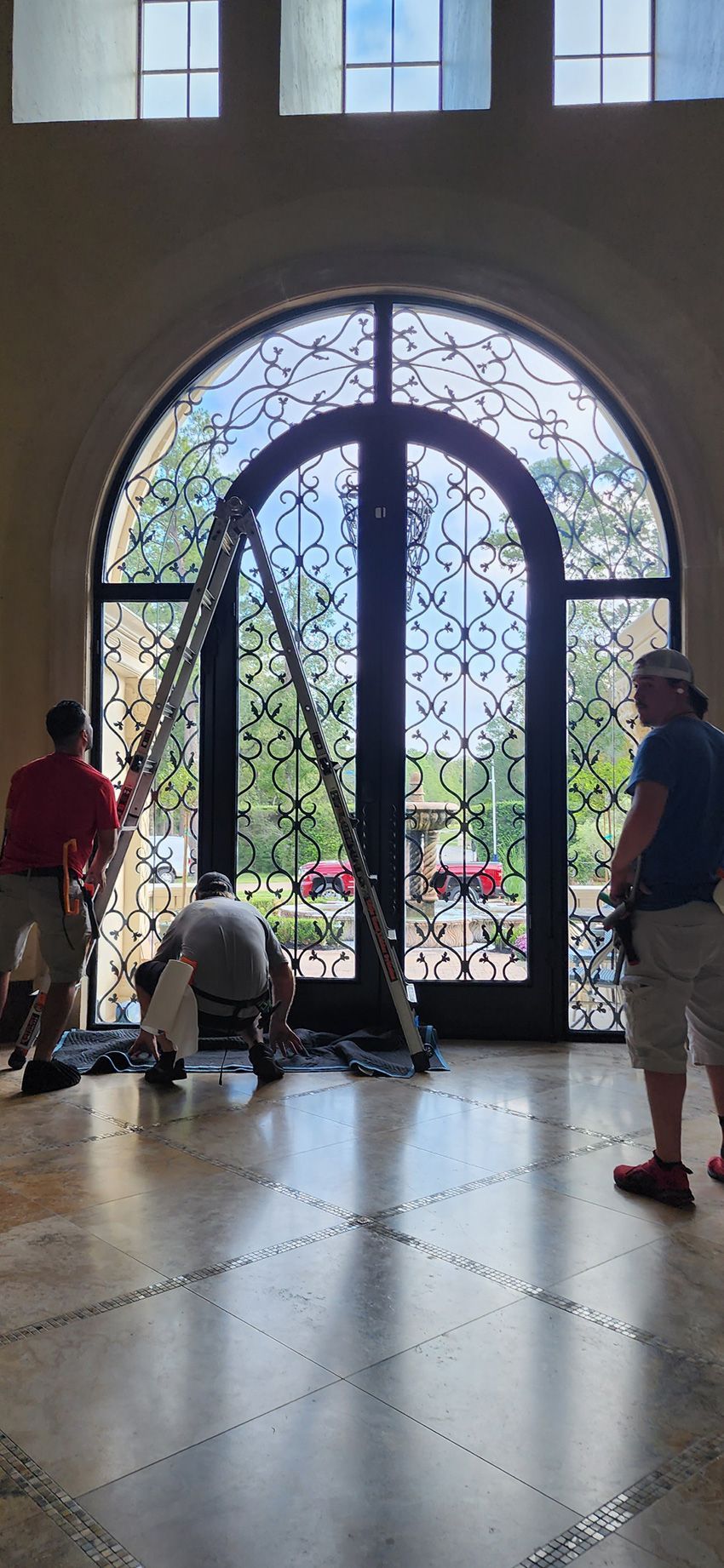 Three people working on a large arched, iron-framed glass doorway in an indoor entryway with marble flooring.