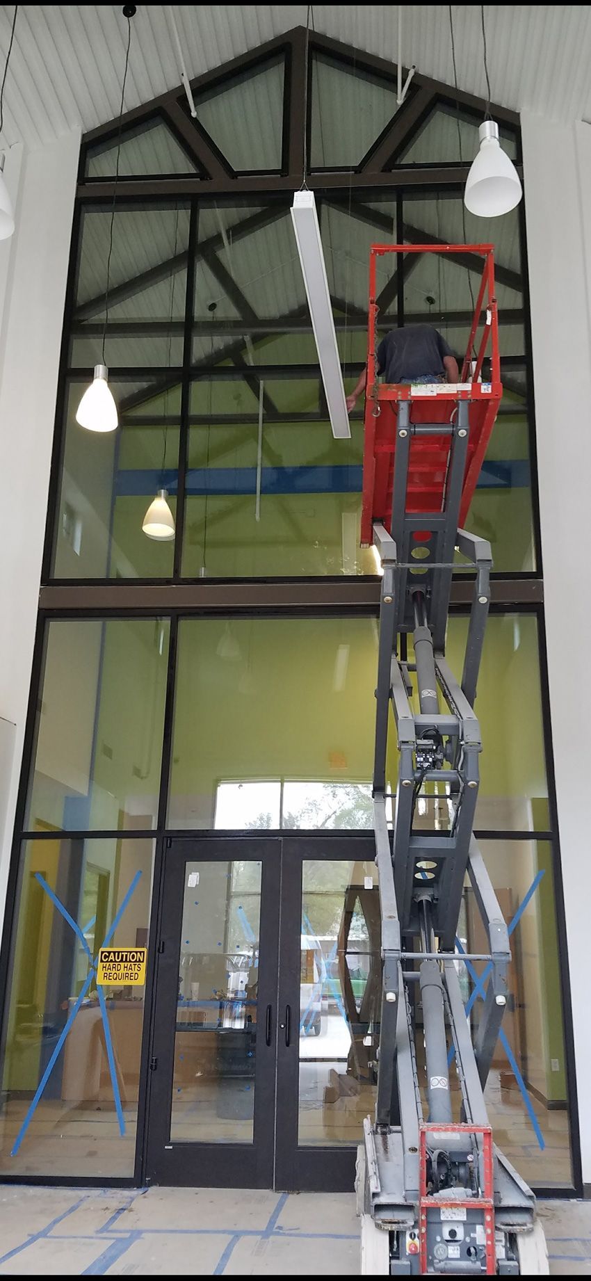 A person in a red scissor lift works on a light fixture installed in front of a tall, glass-paneled building entrance.