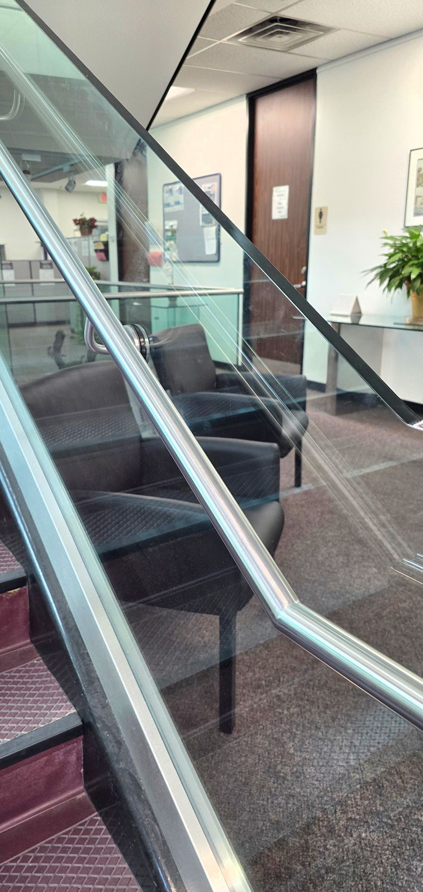 View from a staircase looking through a glass railing at a waiting area with two black chairs and a closed door.