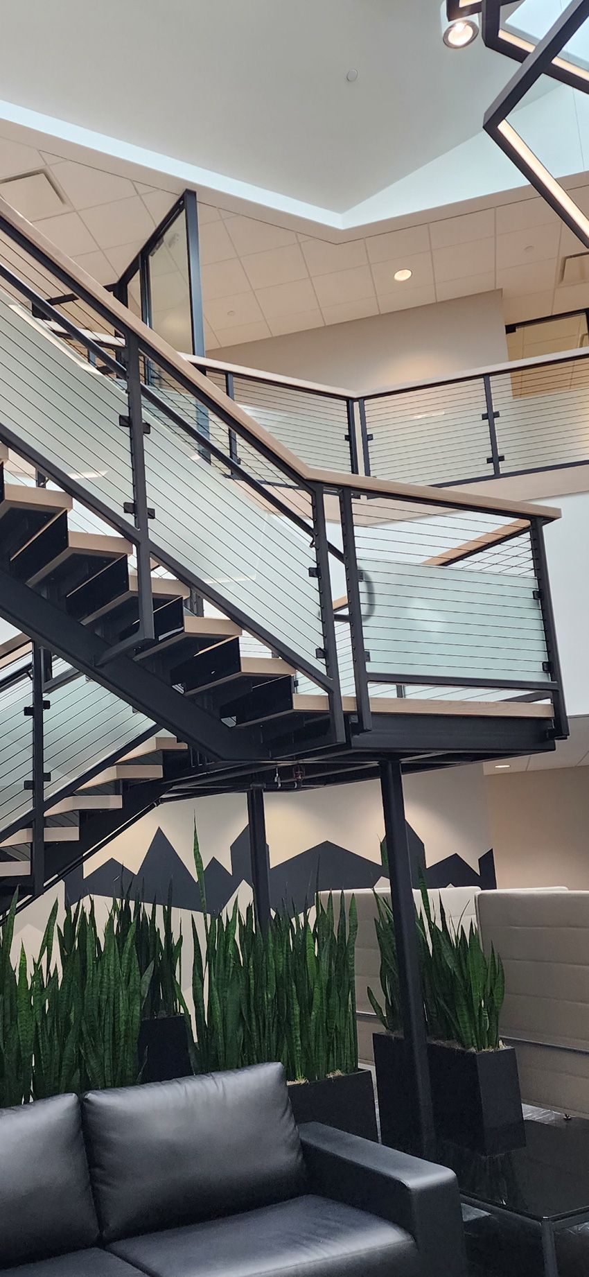 A modern architectural staircase with dark metal railings and glass panels above a sofa and tall indoor plants.
