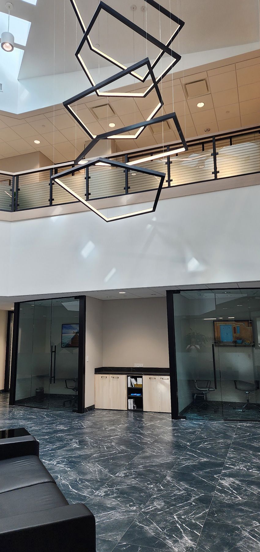 Modern office lobby featuring high ceilings, a dramatic geometric chandelier, glass-walled rooms, and tiled flooring.