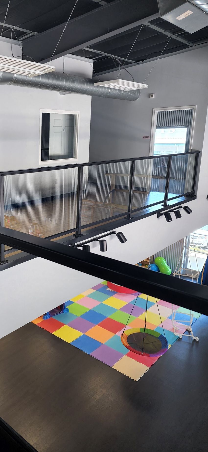 A mezzanine overlooks a room with a colorful tiled floor, a hanging saucer swing, and a sleek black metal railing.