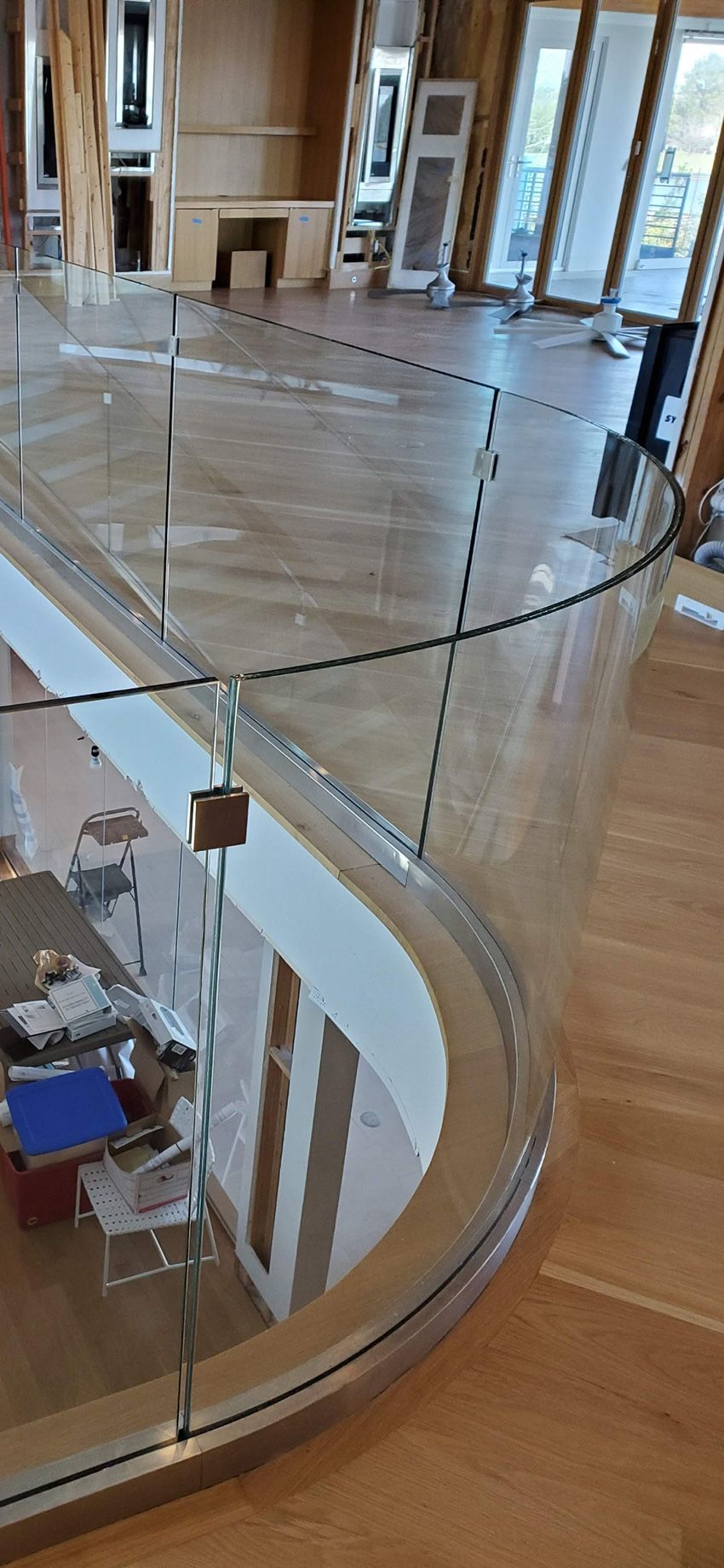 A curved glass railing with a metal base on a wooden floor, overlooking an interior space below.