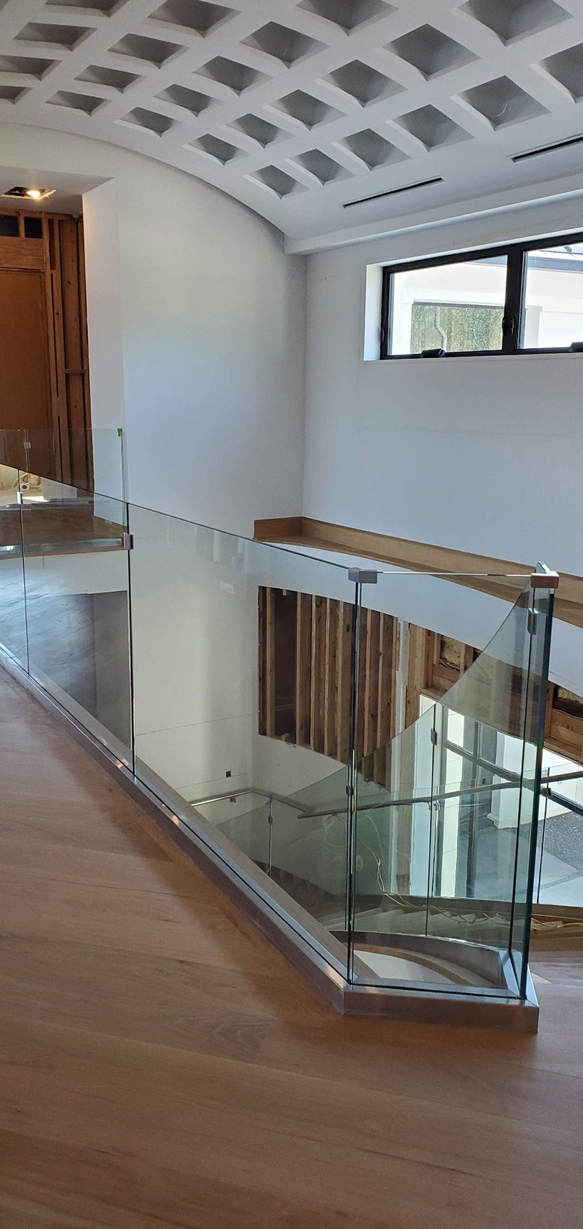 A modern balcony with a glass railing overlooking a wooden staircase, featuring a geometric coffered ceiling above.