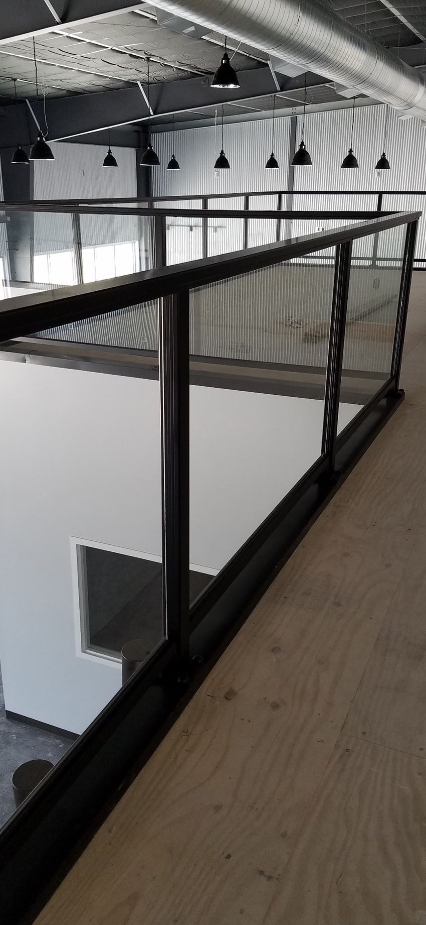 A black metal railing overlooks a high-ceilinged industrial space with hanging pendant lights and a plywood floor.