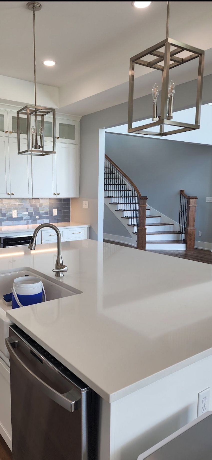 A modern white kitchen island with a sink and metal pendant lights, overlooking a living space with a wooden staircase.